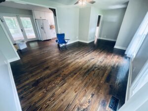 A room with worn pine wood floors, showing dark stains and scratches, white kitchen appliances, and large windows with a view of a porch.